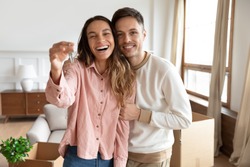Happy young family couple holding key to new home on moving day concept, first time real estate owners man husband embrace woman wife look at camera proud buying property stand in own flat with boxes