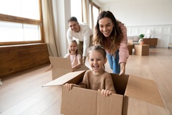 Happy parents playing with cute small kids daughters laughing on moving day, family tenants renters homeowners and children girls having fun riding in box in living room relocating new home concept