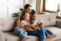 Happy family young mother babysitter hold read book relax embrace cute little children daughters, smiling parent mum tell small kids funny fairy tale story sit on sofa having fun together at home