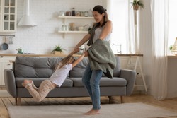 Happy young mother lifting spinning carefree small child daughter hold hands flying in air dancing in modern kitchen, active playful mom having fun with cute little kid enjoy playing together at home