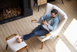 Relaxed casual guy lounge sit on comfortable armchair in modern house room with wooden floor fireplace using smartphone apps, happy young man hold cell phone play mobile games at cozy home, top view