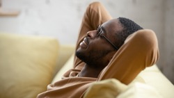 Happy african American young man in glasses sit relax on cozy couch lean hands over head taking nap in living room, smiling biracial male rest on sofa at home sleeping or dreaming, stress free concept