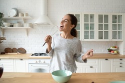 Happy young woman holding beater microphone singing song dancing cooking alone in modern kitchen, funny lady housewife having fun listening music prepare healthy morning meal doing housework at home