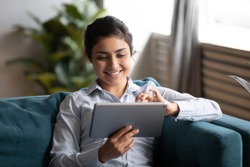 Happy young adult indian woman hold digital pad tablet computer sit on sofa at home, smiling lady using app reading electronic book relax in living room enjoy browsing web on modern device technology