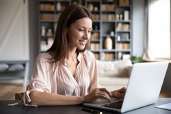 Happy young woman sit at desk look at laptop screen browsing surfing wireless Internet at home, smiling millennial female employee work on modern computer in living room, technology concept