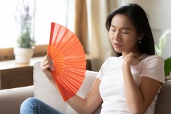 Unwell young Asian woman sit on couch wave with hand fan feel sick at home, overheated millennial Vietnamese girl relax on sofa in living room breathe fresh air from waver, suffer from heatstroke