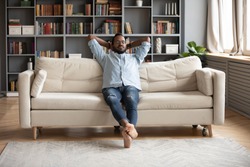 Serene relaxed barefoot young african man resting on comfortable couch in modern living room holding hands behind head, millennial hipster guy enjoy no stress peace of mind lounge on sofa at home