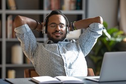 Relaxed african business man wear wireless headphones enjoy listen music with eyes closed hand behind head sit at work desk, young worker take break in office feel peace of mind stress relief concept