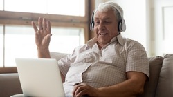 Smiling senior man wear earphones wave to camera having video call on laptop, happy elderly male in headphones sit on couch at home talk using modern technologies and wireless connection