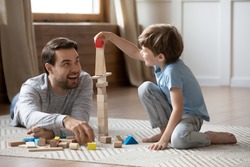 Full length cheerful young man lying on floor carpet, constructing building with wooden blocks with little son. Happy father playing with small schoolboy in living room, enjoying weekend time.