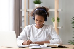 Focused african american teenage girl wearing headphones writing notes study with laptop and books, serious black female high school teen student listening audio course or music while doing homework