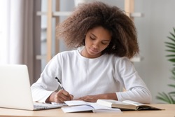 Focused african american school girl studying with books laptop preparing for test exam writing essay doing homework at home, teenage student learning assignment making notes, teen education concept