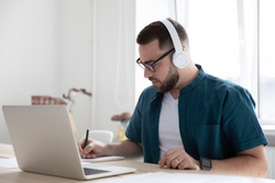 Focused young man businessman company worker employee in glasses wearing wireless headphones, watching educational webinar lecture seminar on laptop online, writing down notes in modern office.