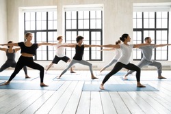 Group of multiracial people practicing yoga asanas, doing Warrior Two Virabhadrasana 2, work out indoors full length, posture increases stamina and flexibility, improves physical and mental endurance