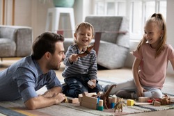 Happy little boy playing with toy dinosaur, having fun with smiling father and elder cute sister in living room. Joyful father enjoying playtime with small children siblings, lying on floor carpet.