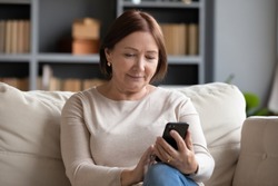 Pleasant senior older lady resting on couch, using applications on smartphone. Happy old mature woman chatting in messenger or social network with friends or children, shopping in internet store.