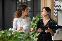 Diverse employees chatting during coffee break, walking in modern office, Asian businesswoman wearing glasses sharing ideas, discussing project with colleague, having pleasant conversation