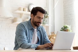 Focused young man wearing glasses using laptop, typing on keyboard, writing email or message, chatting, shopping, successful freelancer working online on computer, sitting in modern kitchen