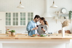 Happy loving couple holding wine glasses, touching foreheads, enjoying tender moment, romantic date, standing in modern kitchen at home together, smiling wife and husband celebrating anniversary