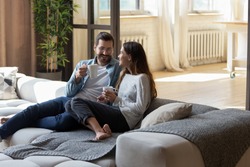 Loving couple relaxing on cozy sofa in living room, drinking hot beverages, tea or coffee, happy smiling man and woman holding white cups sitting on couch, chatting, talking, spending lazy weekend