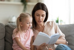 Smart little preschooler girl sit on couch with young mother or nanny reading interesting book at home, small daughter sturdy learn with mom at home enjoy fairytale spend leisure time together