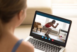 Focus on laptop screen with young woman in sportswear doing morning exercises, deep squats on yoga mat, staying fit at home. Interested girl watching online educational fitness workshop training.