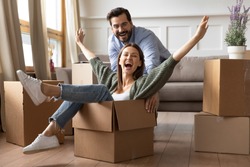 Full length overjoyed young bearded man in glasses pushing laughing wife in carton box. Energetic happy woman sitting in carboard container, having fun with smiling husband in new apartment house.