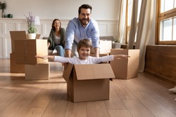 Full length happy young father in glasses pushing carton box with small kid son in new living room. Overjoyed family couple having fun with little child, celebrating moving into flat apartment.