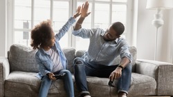 African young father and daughter play enjoy video games sit on couch, giving high five celebrating successful finish or start competition, activity with kids, fun concept