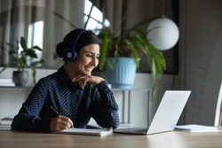 Happy young indian girl with wireless headphones looking at laptop screen, reading listening online courses, studying remotely from home due to pandemic corona virus world outbreak, quarantine time.