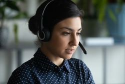 Head shot close up view young serious focused smart attractive indian ethnicity woman wearing modern wireless headset with microphone, working remotely online, consulting client by video call.