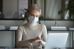 Front view 30s businesswoman wearing protective medical facemask sitting at table with laptop, disinfecting hands before starting working remotely from home, healthcare habit stop spreading virus.