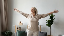 Excited elderly mature retired woman dancing in living room with widely opened outstretched arms, enjoying freedom. Overjoyed happy older female pensioner satisfied with leisure weekend time at home.