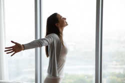 Happy young woman stand near window breathe fresh air stretch exercise in bedroom, smiling millennial girl overjoyed welcome new sunny morning at home or hotel, optimism, happiness concept