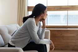 Unhappy young african american girl sit on sofa at home and looking at window, thinking about problem and trouble, suffering from depression or loneliness, mental illness, makes difficult decisions