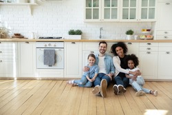 Portrait of happy multiracial young family with little kids sit on warm floor in design modern kitchen, smiling multiethnic parents with small biracial daughters relax in new renovated home together