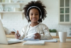 Portrait of smiling little biracial girl in headphones do homework study online in kitchen, happy small African American child in earphones have online web class or lesson using laptop at home