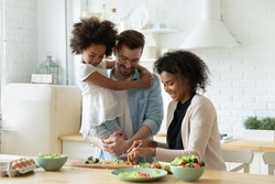 Happy young diverse parents have fun teach little biracial daughter cooking, overjoyed multiracial family with small girl child preparing food making healthy salad for breakfast in kitchen together
