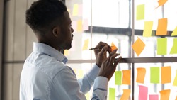 Focused African American male employee write on colorful sticky notes develop business project in office, concentrated biracial man worker brainstorm engaged in creative thinking make startup plan
