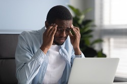 Unhappy african american businessman suffering from headache at work at laptop. Diverse male employee with eyewear stressful touching temples thinking about business problem.
