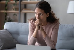 Nervous young woman sitting on sofa, worrying about bad news, received by email on laptop. Stressed businesswoman thinking of crisis challenges, feeling desperate or unsure, stack with hard decision.