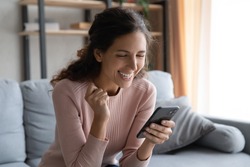 Joyful millennial woman sitting on couch, reading sms with good luck news. Happy young lady celebrating online lottery win, enjoying communicating with friends or beloved man, feeling excited at home.