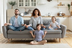 Full length peaceful calm couple practicing yoga exercises with small children in studio living room. Mindful little boy sitting on floor in lotus pose while parents relaxing on sofa with sister.