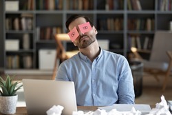 Exhausted tired businessman with painted eyes on stickers, adhesive notes on face sleeping at workplace, sitting at desk with laptop, unproductive lazy young male dozing, working on difficult project