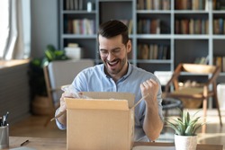 Excited happy man looking into cardboard box, sitting at work desk, received parcel with awaited online store order, smiling laughing young male celebrating success, lottery winner prize