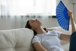 Exhausted young woman sit rest on sofa at home use waver suffering from hot weather indoors, overheated unwell millennial female breathe fresh air, wave with hand fan, having heatstroke