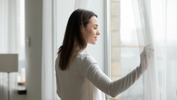 Young woman open curtains at home or hotel look in window distance thinking or dreaming, happy millennial female admire view, feel optimistic and positive, welcome new sunny day, early morning