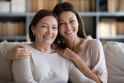 Head shot portrait smiling mature mother and daughter hugging, sitting on couch at home, looking at camera, young woman embracing older mum shoulders, family photo, two generations bonding