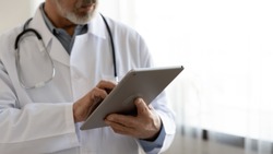 Old male doctor holding modern digital tablet computer standing in hospital, close up view. Senior middle aged professional medic wears white coat and stethoscope using healthcare ehealth application.