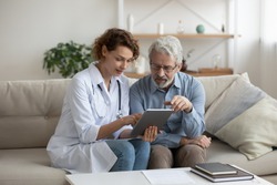 Female professional doctor showing medical test result explaining prescription using digital tablet app visiting senior man patient at home sitting on sofa. Elderly people healthcare tech concept.